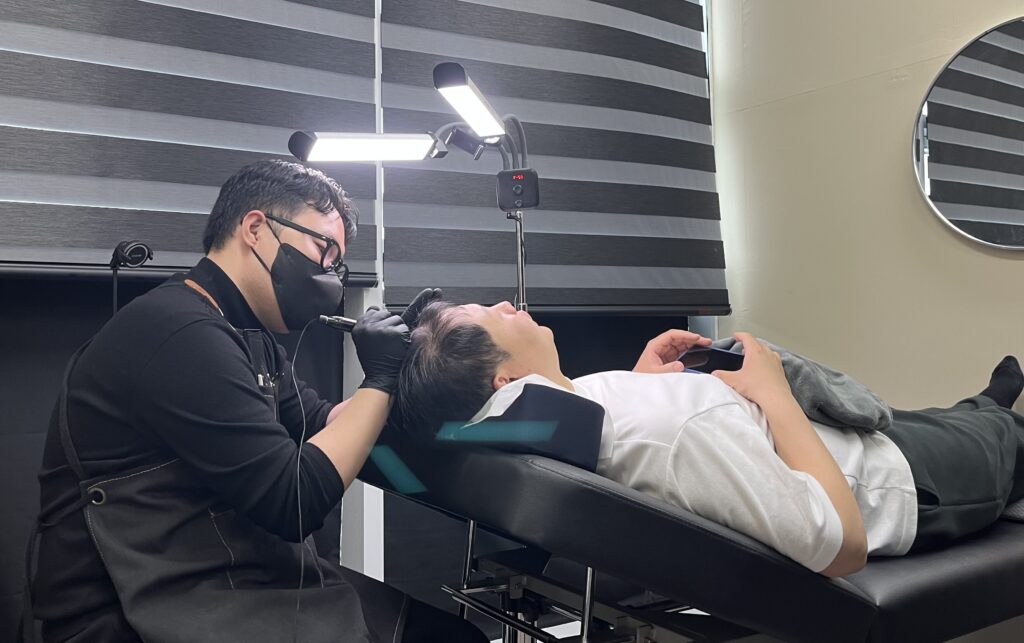 시술 베드에 누운 고객의 헤어라인에 단계적으로 밀도를 올리며 시술하는 모습